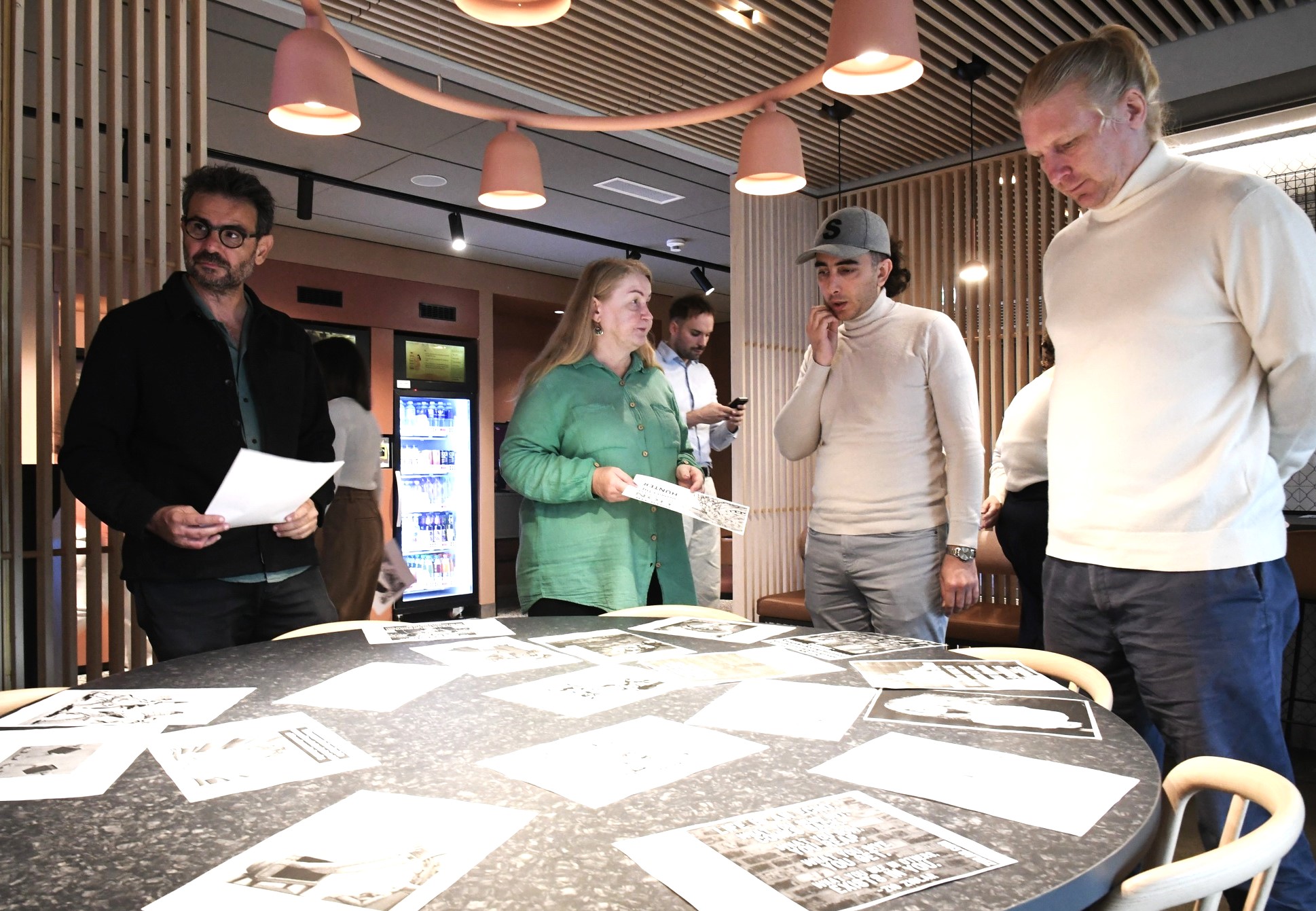 Four participants stand around a round table covered with printed black and white images and handouts, discussing them together during a practical session of the HATE-LESS Training of Trainers in Tallinn.