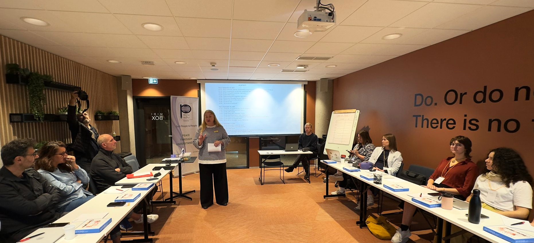 A trainer stands and speaks to participants seated at tables during the opening session of the HATE-LESS Training of Trainers in Tallinn. A roll-up banner beside her carries the People to People International globe emblem and the words Eesti People to People, Peace Through Understanding. The projected screen behind her shows the three-day agenda, beginning on 15 September 2025.