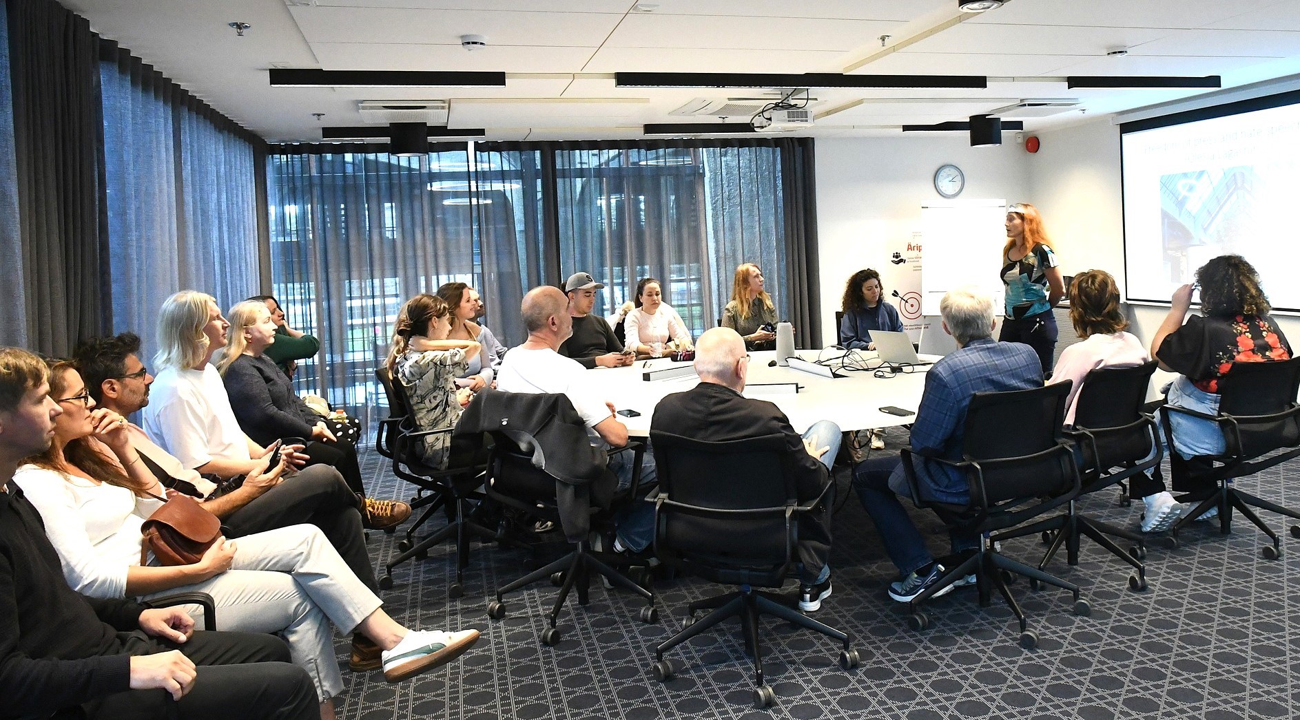 About twenty participants sit around a large table and in chairs along the wall of a bright meeting room in Tallinn, listening to a trainer who is standing and presenting beside a projected slide, during the HATE-LESS Training of Trainers in September 2025.